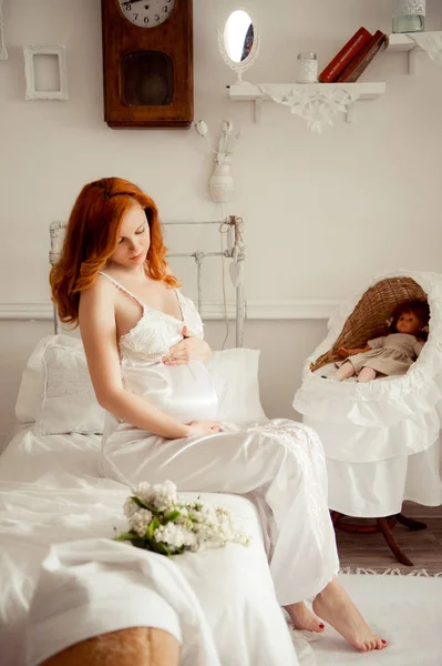 Mujer embarazada con el pelo rojo en vestido blanco en la habitación de los niños — Foto de Stock