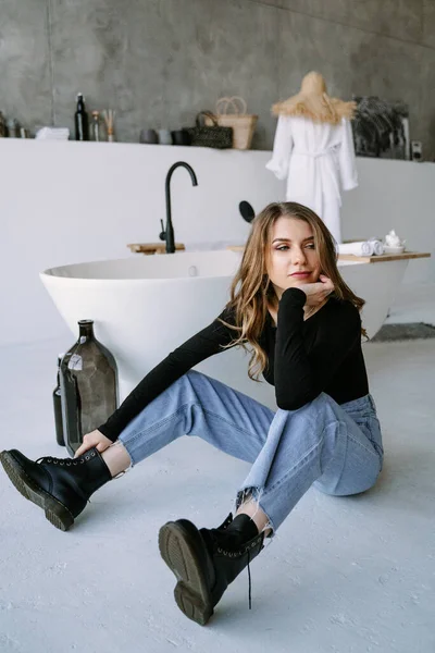 Portrait d'une belle fille aux cheveux longs marron clair en t-shirt noir, jean et bottes rugueuses. Salle de bain élégante — Photo