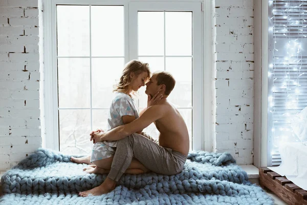 Stylish interior. A couple near the window, under one blue blanket of large mating. Gentle embrace and kisses. Blonde. Floor lamp. Athletic build. Gray pajamas in hearts. Love story.