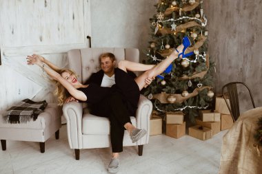 Bir adam ve bir kız yılbaşı ağacının yanında. Siyah bir gece elbisesi ve dudaklarında kırmızı ruj olan sarışın. Bej sandalye. Mavi sandaletler. Noel ağacının altındaki hediyeler. Beyaz ahşap kapılar.