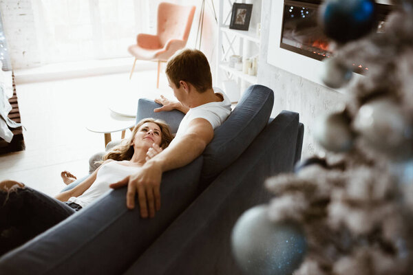 The guy and the girl hug and kiss on the blue couch. Christmas tree decorated with toys. Electronic fireplace. Bright interior. Blonde in a white T-shirt and jeans.