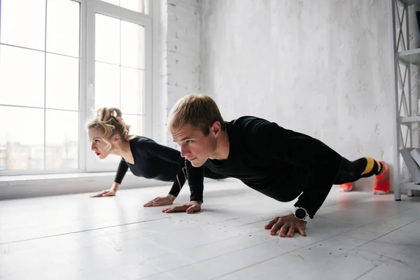 The guy and the girl in tracksuit. Black sports uniform. Male and female athletes. Pumped up body. Push ups. Morning work-out. Set of exercises for body. Classes in pair. Workout together at home.