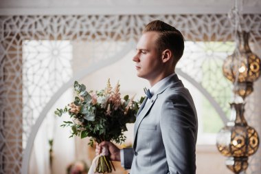 Damat, Fas tarzı bir odada gri takım elbiseli gelini bekliyor. Ellerinde şık bir düğün buketi.