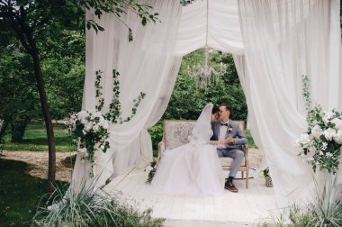 Gelin ve damat bahçede güzel bir kanepede oturuyorlar. Beyaz çadır. Yeşil yapraklar. Pembe kabarık elbiseli gelin, gri takım elbiseli damat..