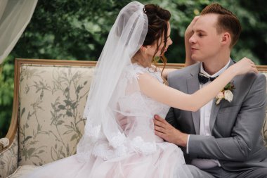 Gelin ve damat bahçede güzel bir kanepede oturuyorlar. Yeşil yapraklar. Pembe kabarık elbiseli gelin, gri takım elbiseli damat..