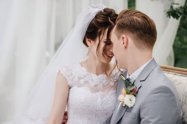 Gelin ve damat bahçede güzel bir kanepede oturuyorlar ve öpüşüyorlar. Yakın bir portre. Beyaz çadır. Yeşil yapraklar. Pembe kabarık elbiseli gelin, gri takım elbiseli damat..