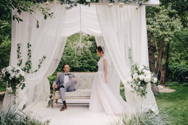 Gelin ve damat bahçede güzel bir kanepede oturuyorlar. Beyaz çadır. Yeşil yapraklar. Pembe kabarık elbiseli gelin, gri takım elbiseli damat..