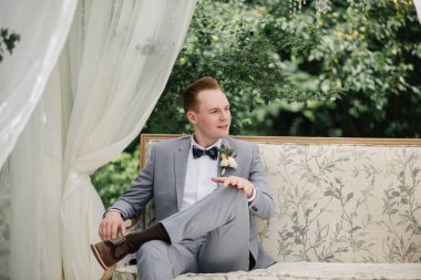 Damat bahçedeki bir çardakta güzel bir kanepede oturuyor. Beyaz çadır. Yeşil yapraklar. Pembe kabarık elbiseli gelin, gri takım elbiseli damat..