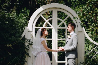 Gelin ve damat arkaplanda beyaz bir kemer üzerinde dururlar. Ormanda düğün töreni. El ele tutuşup birbirlerine bakıyorlar..