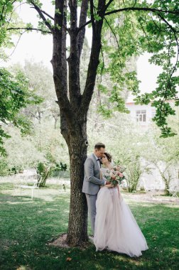 Gelin ve damat bahçede yürüyorlar. Ormanda düğün. Pembe, gür elbise ve gri takım elbise, papyon.