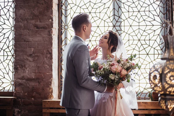 The first meeting. The groom comes into the room to the bride with a bouquet. They hug and kiss. Pink wedding dress, gray suit and stylish bouquet. Room in the style of Morocco.