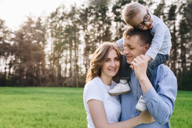 Ormanda piknikte bir aile. Açık bir alanda, yeşil çimlerde oturmak. Mavi kıyafetler. Anne ve baba oğullarıyla oynar, sarılır ve gülümserler. Gözlüklü bir çocuk..