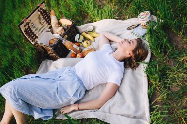 Ormanda piknikte bir kız. Ormanlık alanda, yeşil çimlerde oturuyor. Mavi kıyafetler. İçinde yiyecek olan piknik sepeti. Doğada öğle yemeği. Sarışın, kısa saçlı. Modaya uygun yıkama.