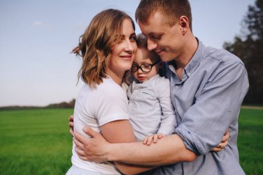 Ormanda yürüyüş yapan bir aile. Çayırda piknik, yeşil çimlerin üzerinde. Mavi kıyafetler. Anne, baba, oğul. Oğul babanın ellerinde. Evcil hayvan. Kara Köpek. Gözlüklü çocuk..