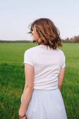 Kız sarışın, kahverengi saçlı, beyaz gömlekli ve mavi etekli. Yeşil çimlerin arasında, tarlada yürüyorum. Bir kızın portresi. Olumlu ve gülümse..
