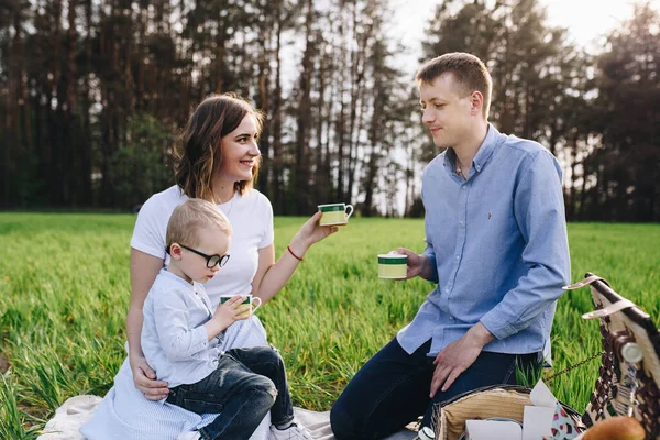 Family in the forest at a picnic. Sit in a clearing, green grass. Blue clothes. Mom and Dad play with their son, hug and smile.