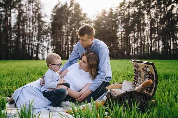 Family in the forest at a picnic. Sit in a clearing, green grass. Blue clothes. Mom and Dad play with their son, hug and smile. A child with glasses.