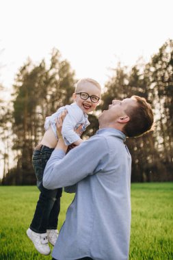 Aile içinde bir aile. Ormanda, çayırda piknik. Yeşil çimenler. Mavi kıyafetler. Baba, gözlüklü oğlum. Sarı saçlı bir çocuk. Neşe. Babam oğlunu havaya fırlattı. Birlikte.