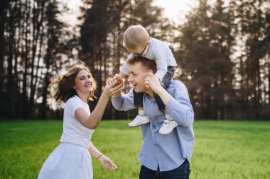 Aile içinde bir aile. Ormanda, çayırda piknik. Yeşil çimenler. Mavi kıyafetler. Anne, baba, gözlüklü oğul. Sarı saçlı çocuk. Neşe. Ebeveynler çocukla oynarlar. Birlikte. Piknik sepeti. Yemek, battaniye