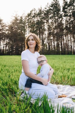 Ormanda piknikte bir aile. Açık bir alanda, yeşil çimlerde oturmak. Mavi kıyafetler. Anne oğluna sarılıyor. Gözlüklü bir çocuk. Birlikte zaman geçirmek