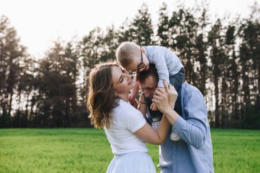Ormanda piknikte bir aile. Açık bir alanda, yeşil çimlerde oturmak. Mavi kıyafetler. Anne ve baba oğullarıyla oynar, sarılır ve gülümserler. Gözlüklü bir çocuk..