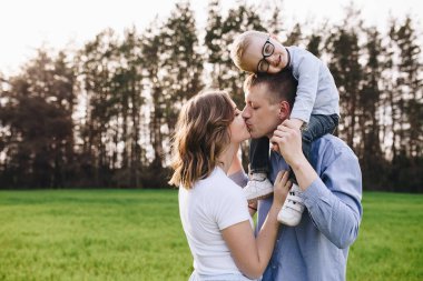 Ormanda piknikte bir aile. Açık bir alanda, yeşil çimlerde oturmak. Mavi kıyafetler. Anne ve baba oğullarıyla oynar, sarılır ve gülümserler. Gözlüklü bir çocuk..