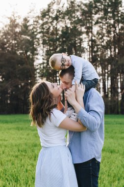 Ormanda piknikte bir aile. Açık bir alanda, yeşil çimlerde oturmak. Mavi kıyafetler. Anne ve baba oğullarıyla oynar, sarılır ve gülümserler. Gözlüklü bir çocuk..