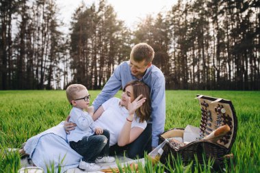 Ormanda piknikte bir aile. Açık bir alanda, yeşil çimlerde oturmak. Mavi kıyafetler. Anne ve baba oğullarıyla oynar, sarılır ve gülümserler. Gözlüklü bir çocuk..