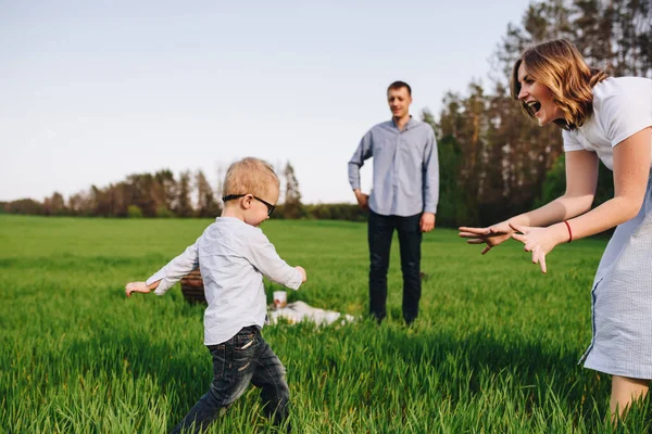 Family in nature. Picnic in the forest, in the meadow. Green grass. Blue clothes. Mom, dad, son with glasses. Boy with blond hair. Joy. Parents play with child. Together. Picnic basket. Food, blanket