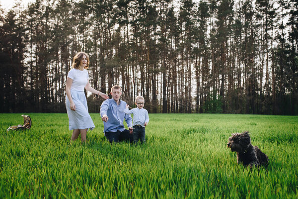 Family in the forest at a picnic. Sit in a clearing, green grass. Blue clothes. Mom and Dad play with their son, hug and smile. A child with glasses. Time together Picnic basket with food. Pet - dog.