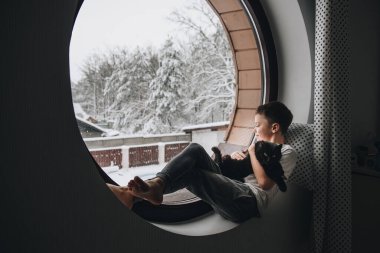 Pencerede pijamalı çocuk. Elinde siyah bir kedi tutuyor. Yuvarlak pencereden güzel bir manzara. Ev rahatlığı. İç mekan. Evin yerleşim planı..