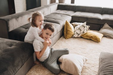 Büyük bir evde aile. Yaşam tarzı ev konforu. Evdeki çocuklar. Yerde oynayan çocuklar. Tekstil. Erkek ve kız kardeş kucaklaşması. Kahverengi kanepe, sarı yastıklar.