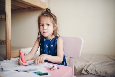 Masadaki çocuk odasında koyu renk saçlı bir kız keçeli kalemle resim yapıyor..