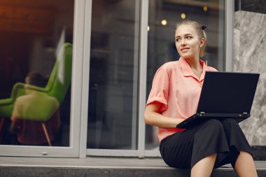 Pembe tişörtlü kız evin yanında oturuyor ve bilgisayarı kullanıyor.