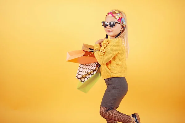 Cute little girl with shopping bags on a yellow background — Stock fotografie