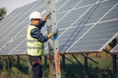Güneş panelinin yanında beyaz kasklı bir adam var.
