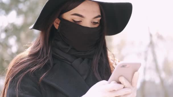 Chica joven en una máscara y guantes está utilizando su teléfono inteligente en un parque — Vídeo de stock