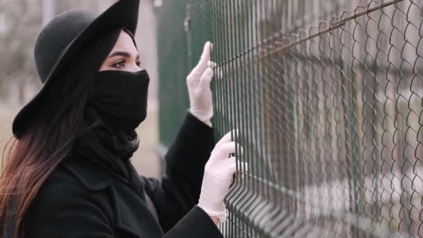 Mooie vrouw in een zwart beschermend masker kijkt door een hek — Stockvideo