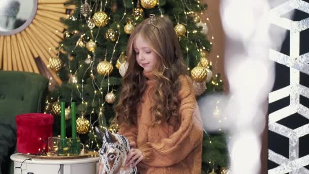 Mała dziewczynka siedzi na koniu zabawki w pobliżu choinki — Wideo stockowe