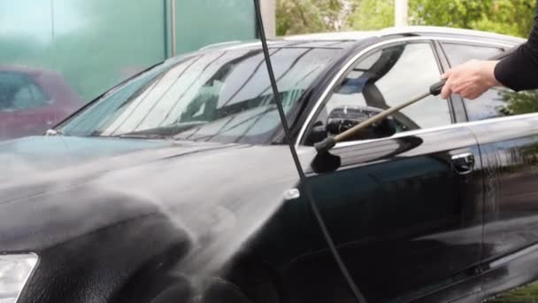 El hombre está enjuagando un coche negro en un punto de auto-lavado con la varita pulverizadora — Vídeos de Stock