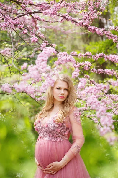Hermosa mujer embarazada en vestido suavemente rosa y tocando el vientre está de pie cerca de la flor de cerezo — Foto de Stock