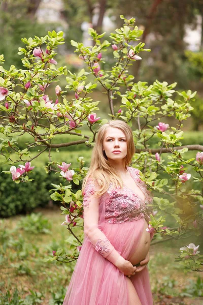 Young future mother in achic pink dress is standing near the blooming magnolia