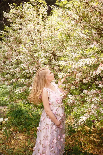 Hermosa mujer embarazada en vestido de encaje rosa tocando el vientre y oliendo flores en el árbol Imagen De Stock