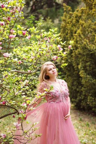 Madre embarazada en negligee suavemente rosa se encuentra cerca de la magnolia que florece con flores de color rosa . Imagen De Stock