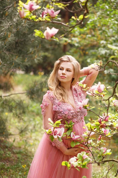 Hermosa mujer embarazada en flores vestido rosa toca el vientre de la mano de pie cerca del árbol de magnolia floreciente Fotos De Stock Sin Royalties Gratis