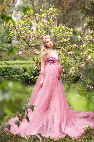 Hermosa joven embarazada en un largo vestido rosa sexy de pie cerca de una magnolia floreciente en la naturaleza Imagen De Stock