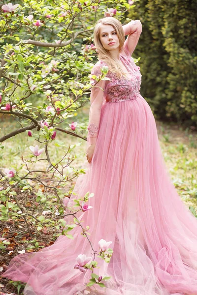 Mujer hermosa joven embarazada en vestido largo de encaje rosa en un jardín con flores Imágenes De Stock Sin Royalties Gratis