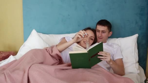 Junges Ehepaar legt sich auf das Bett und liest ein lustiges Buch. — Stockvideo