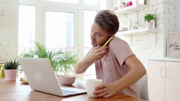 Зайнятий молодий чоловік снідає і використовує свій ноутбук і смартфон — стокове відео
