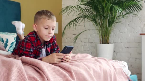 Młody chłopiec od ośmiu lat leżący na łóżku za pomocą smartfona. — Wideo stockowe